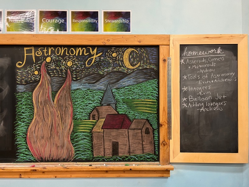 A Waldorf chalkboard astronomy drawing beside a class homework list